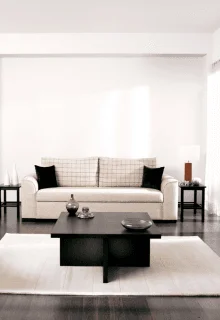 Home 3 A minimalist living room with a white sofa featuring checked cushions and black pillows. A dark coffee table holds decorative items. Flanked by sleek side tables.