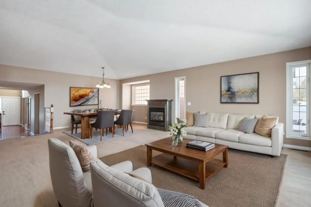 Spacious living room with beige walls, a white sofa, and armchairs. A wooden coffee table with flowers, abstract art, and a cozy dining area in the back.