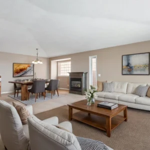 Spacious living room with beige walls, a white sofa, and armchairs. A wooden coffee table with flowers, abstract art, and a cozy dining area in the back.