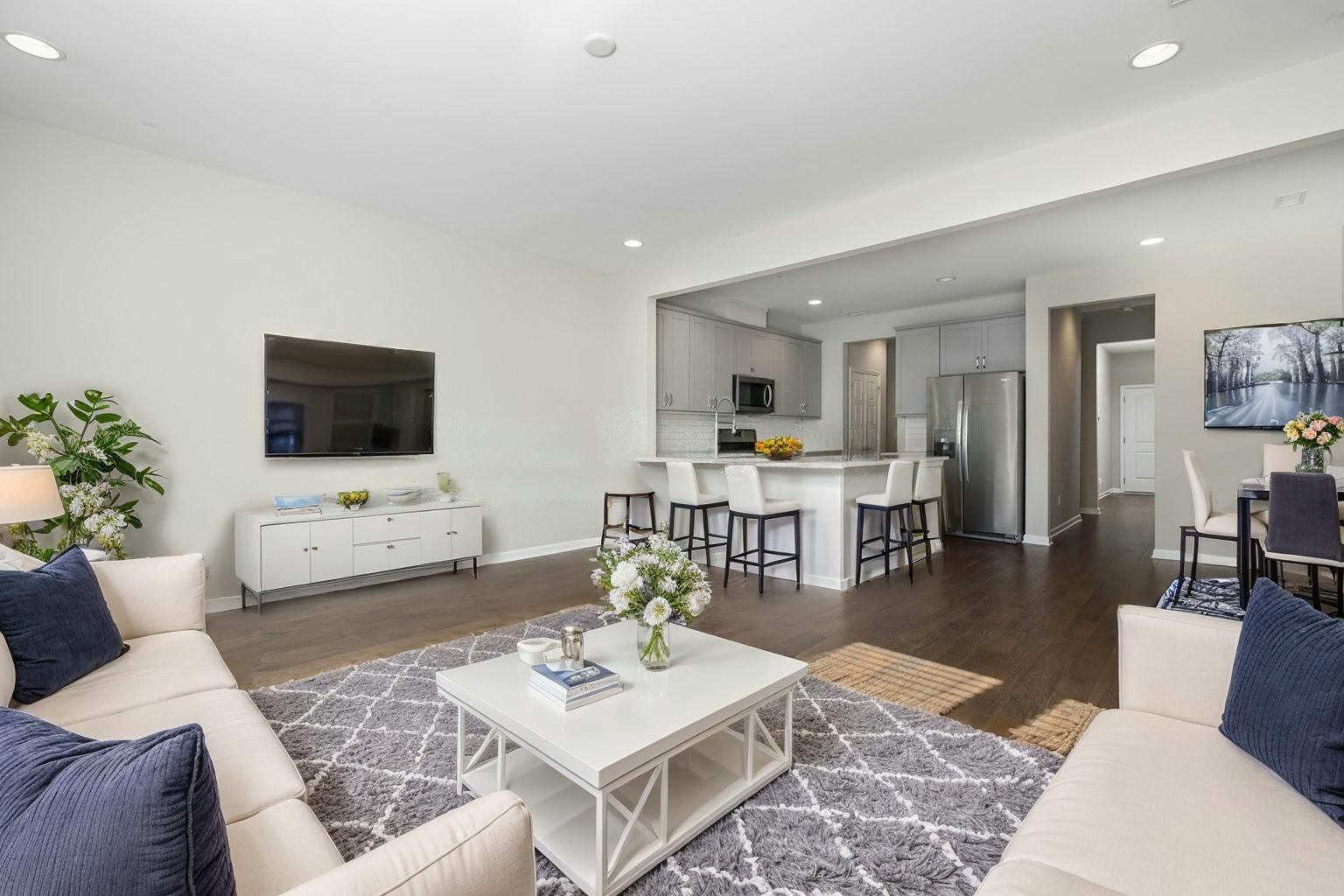Home 2 Modern, open-concept living room and kitchen. Neutral tones, blue accents, large TV, white sofa, island with bar stools, dining area, and wood floors.