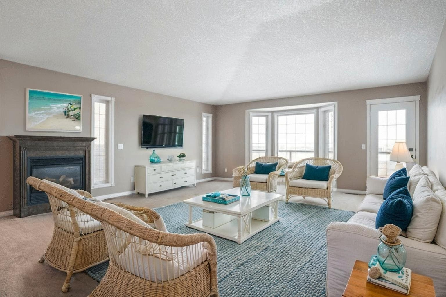Home 17 Spacious, light-filled living room with beige walls, a cozy fireplace, and wicker seating with teal accents. Large windows and a beach-themed decor.