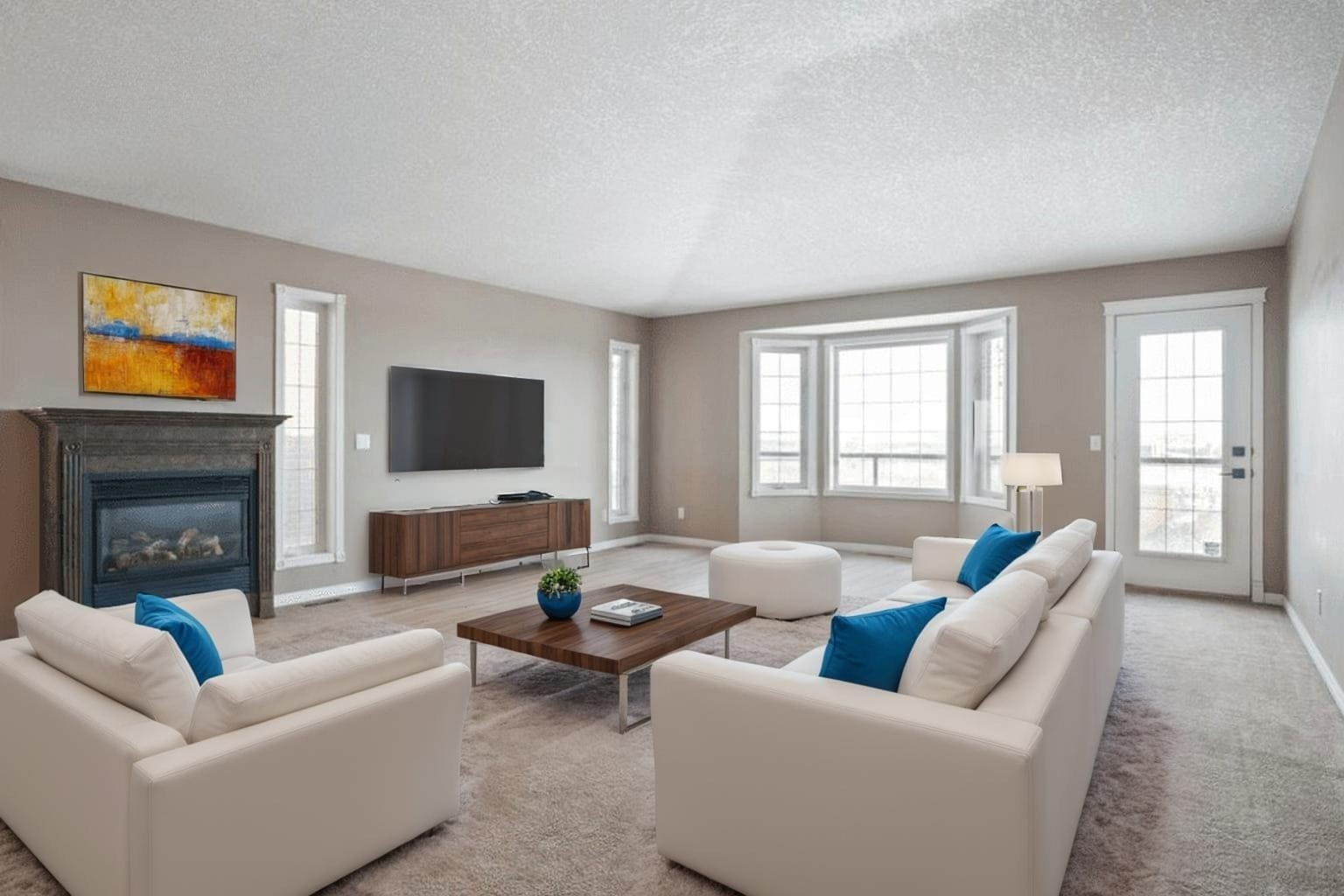 Home 19 Spacious living room with beige walls, white sofas, blue pillows, a wooden coffee table, TV, fireplace, large windows, and a glass door. Cozy ambiance.