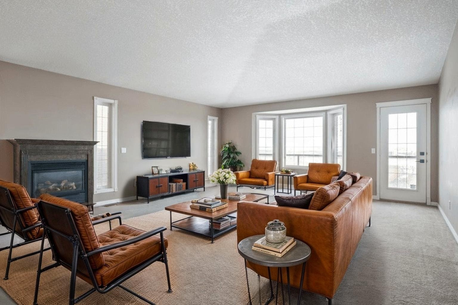 Home 14 Modern living room with a cozy, inviting feel. Features two brown leather sofas, black metal chairs, a fireplace, and a large TV. Ample natural light.