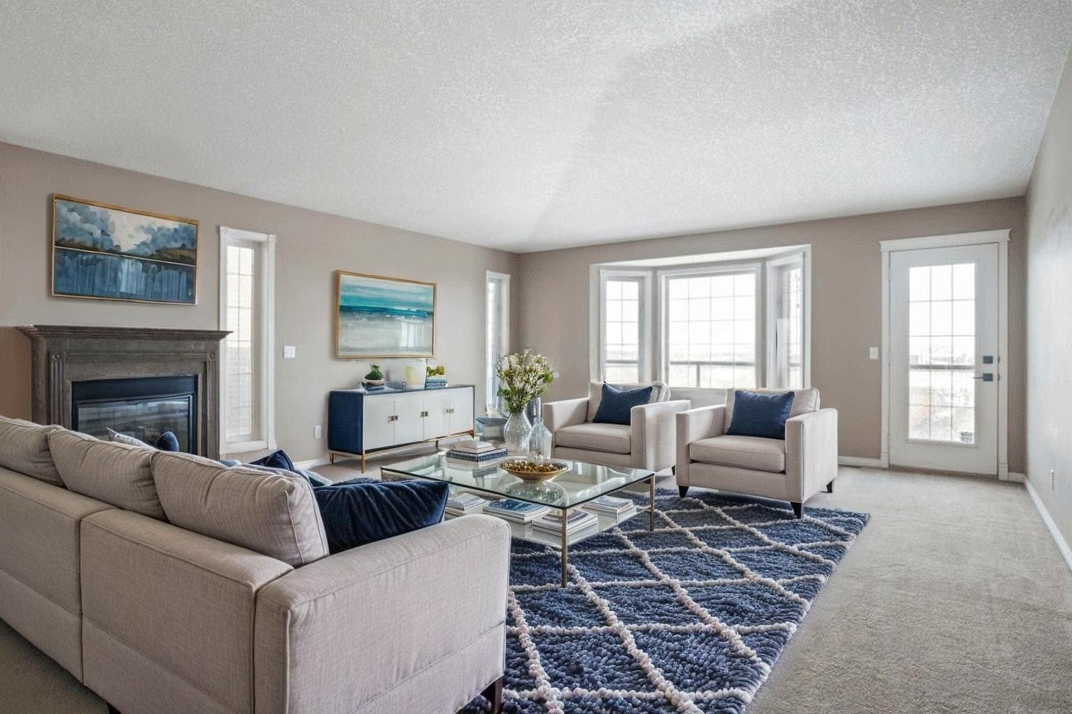 Home 12 Spacious living room with beige walls, featuring a large cream sectional, glass coffee table, and fireplace. Natural light streams through bay windows, enhancing the cozy ambiance. A navy patterned rug and ocean-themed art add elegance.