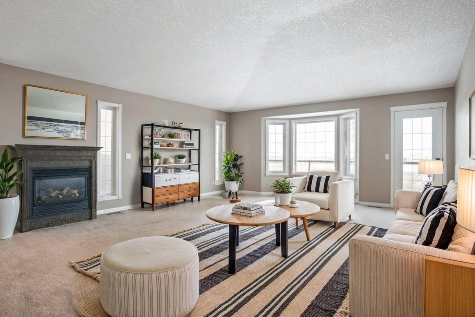 Home 13 A spacious, modern living room with neutral tones. It features a striped rug, round coffee table, cozy seating, plants, a fireplace, and ample natural light.
